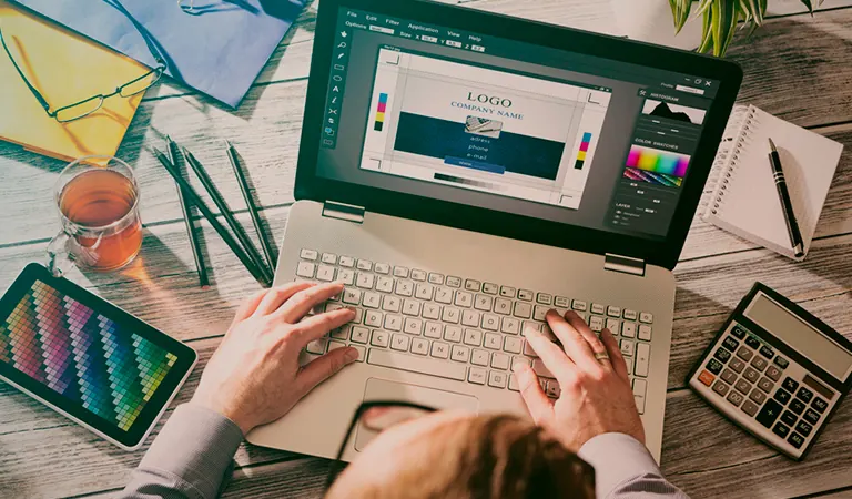 Pessoa criando um logotipo no notebook, ao lado de lápis de cor e calculadora